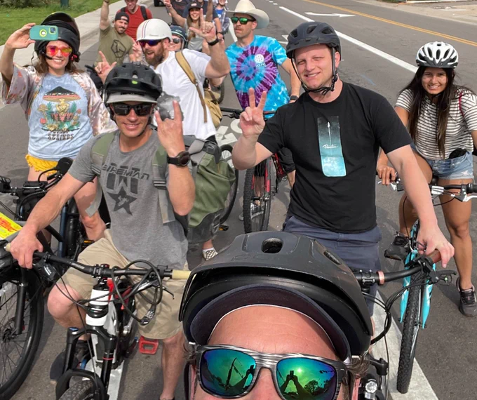 A group of joyful cyclists on a road with their e-bikes, waving and smiling at the camera.