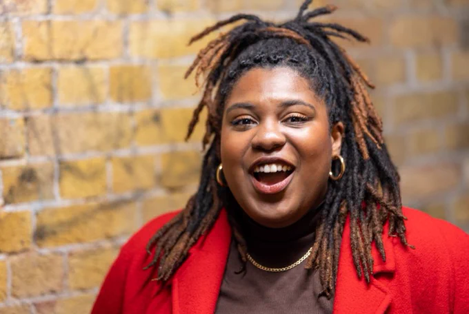 A black femme person with black and brown dreads smiles at the camera. They are wearing a red jacket