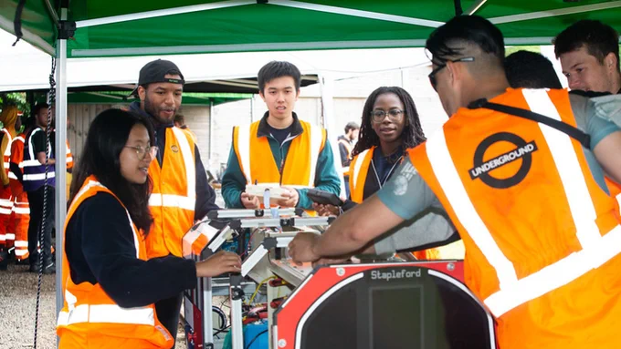 A group of individuals in safety vests, including one with a vest labeled "Underground," collaboratively work around electronic equipment under a tent. One person holds a clipboard.