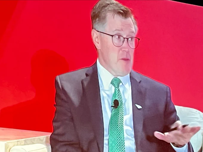 Individual presenting at a conference, seated on stage with a red background, wearing a suit and tie.