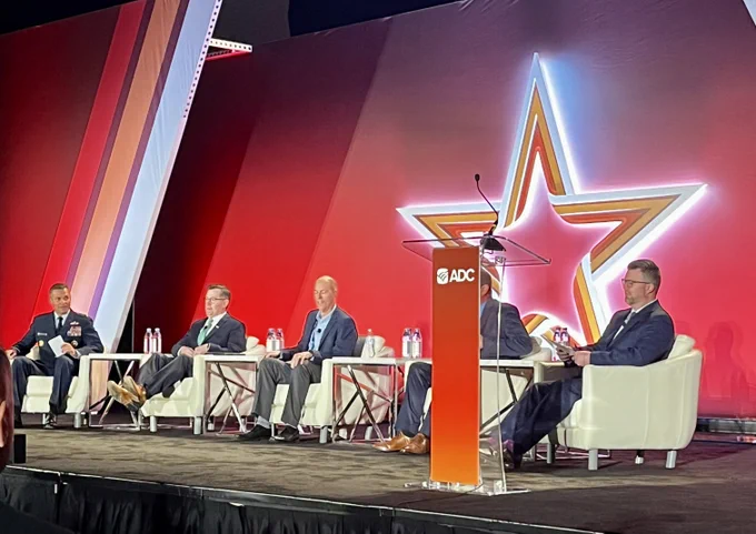 Five panelists, including a military uniformed individual, seated on stage at an ADC event, discussing in front of a large red backdrop with a star logo.