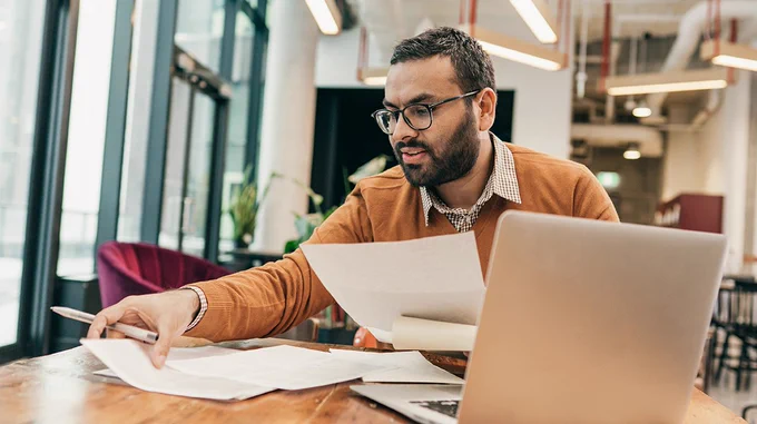 A businessperson sitting at a table in a shared office space looking through papers.