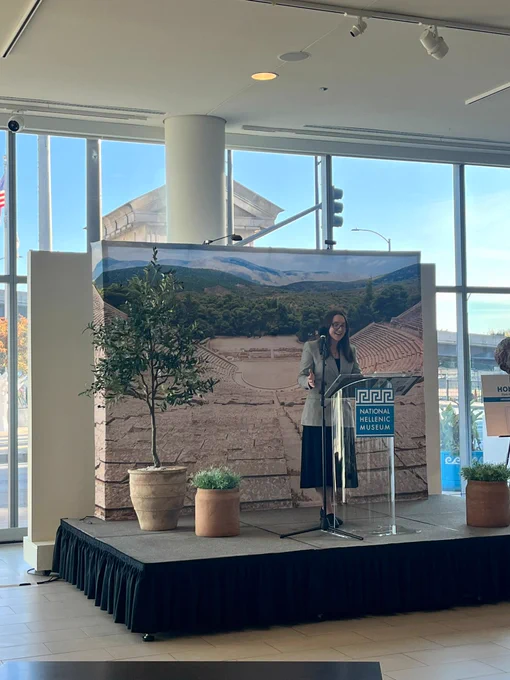Person stands behind podium and gives speech. Greek scenery on tapestry in background.