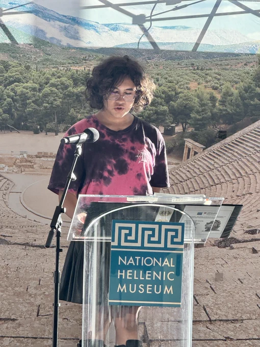 Person stands behind podium and gives speech. Greek scenery on tapestry in background.
