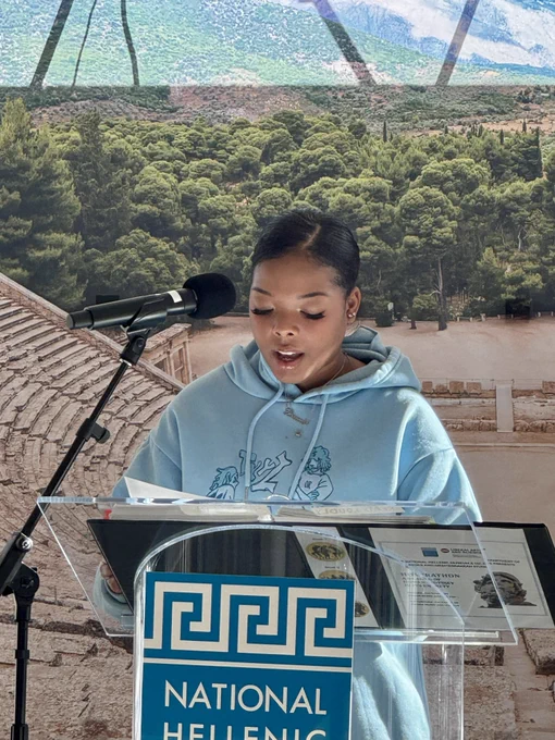 Person stands behind podium and gives speech. Greek scenery on tapestry in background.