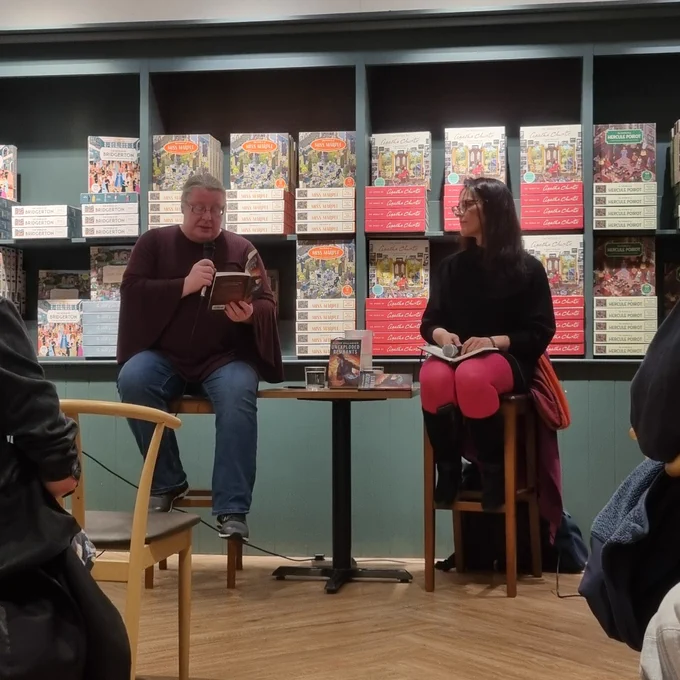 Elaine Gallagher, a grey-haired woman with glasses, reads from a book while holding a microphone. Meg MacDonald, a woman with long black hair and glasses, watches on.