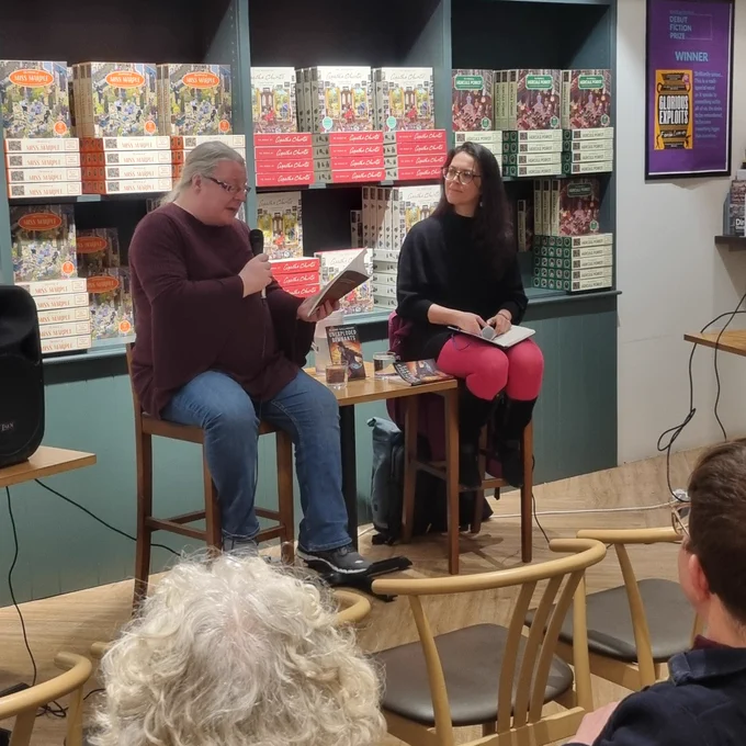 Elaine Gallagher, a grey-haired woman with glasses, reads from a book while holding a microphone. Meg MacDonald, a woman with long black hair and glasses, watches on. 