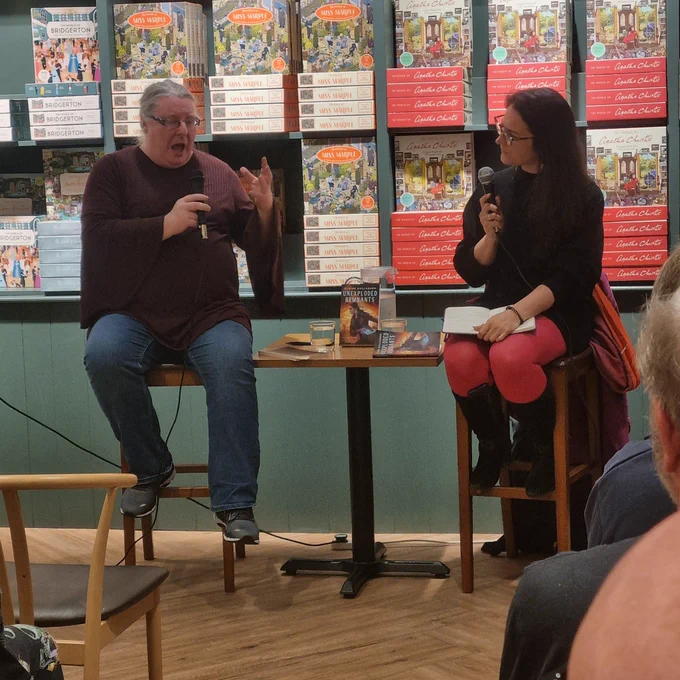 Elaine Gallagher, a grey-haired woman with glasses, gestures and speaks while holding a microphone. Meg MacDonald, a woman with long black hair and glasses, watches on.