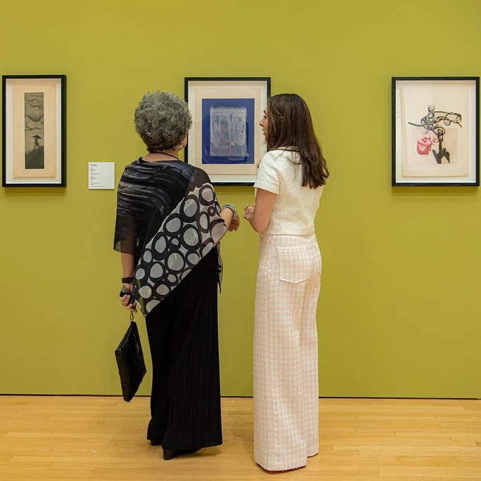 Two individuals viewing and discussing artworks in YBCA's gallery, with three framed pieces by Nahid Hagigat visible on a green wall.