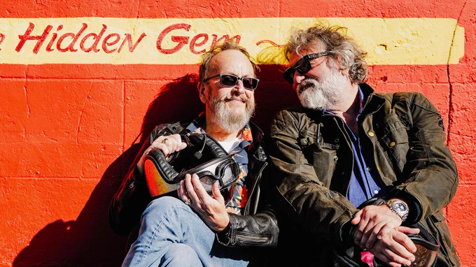 Dave Myers and Si King sitting in their biking gear, leaning against an orange wall and smiling in the sun