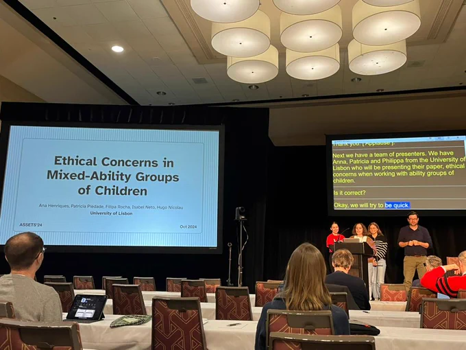 Conference room with slides to the right on Ethical Concerns in Mixed-Ability Groups of Children. To the right, 1 person talking on the podium, 2 behind them, 1 interpreter to their right and captions behind. There are people seated in the chairs before the stage.
