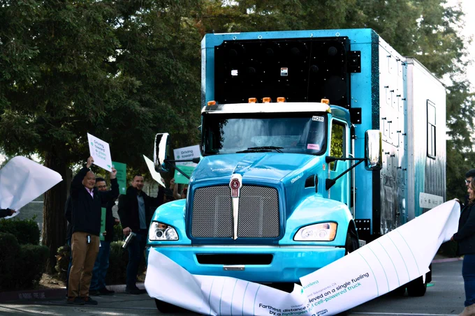 A blue truck driving through a paper banner with people cheering around it.