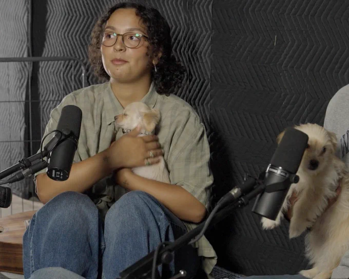 the person who previously had a guitar in their lap now has their knees up to their chest and a sleeping pup held close to their chest. another puppy is hanging over a person’s arm on the right edge of frame.