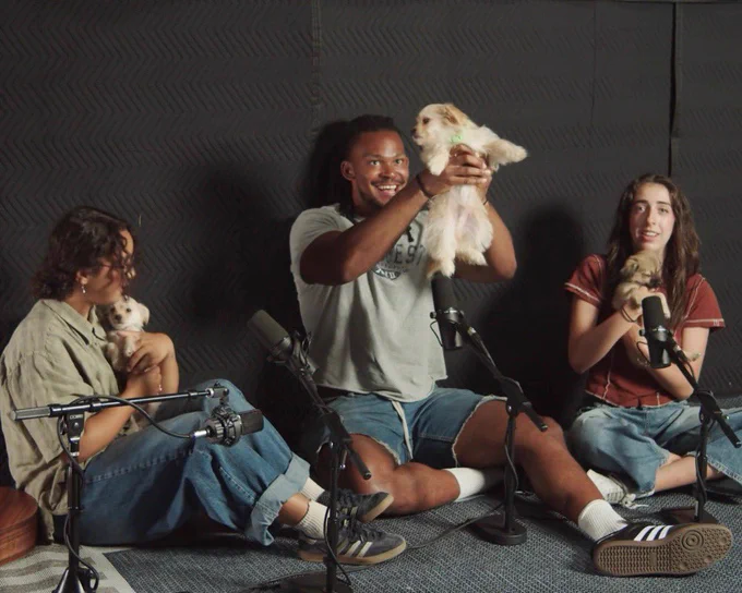 the three members of tiny habits are each holding a puppy. the person in the middle in jean shorts is holding theirs up under the arm pits like the pup is a baby simba cub. the other two pups are being held close to the people’s chests.