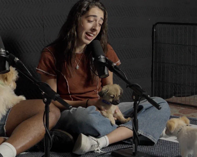 a person in a brown shirt and blue jeans sings into a microphone while holding a puppy in their lap. they’re looking down at a second pup, who appears to be in a staring contest with the first one.