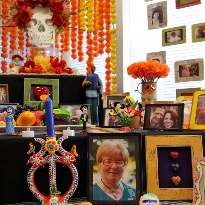 A colorful Day of the Dead altar decorated with marigold flowers, photo frames of various individuals, candles, and decorative skulls. Background features more flowers and figures.