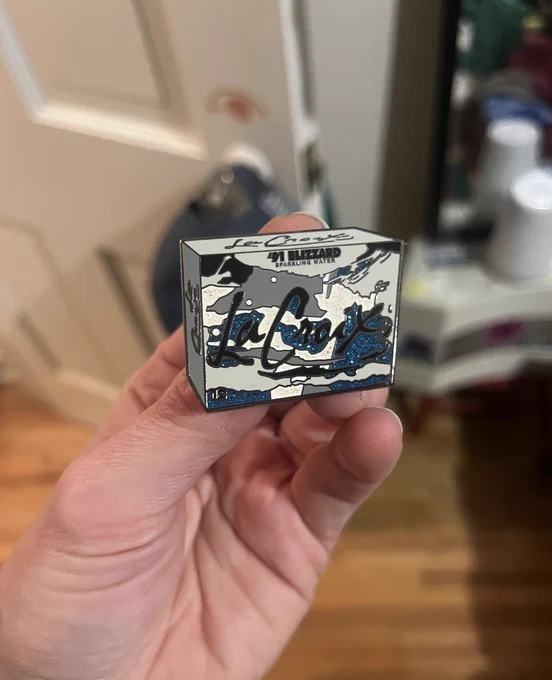 A hand holding up a pin designed as a '91 Blizzard-flavored La Croix 12 pack. The pin is colored in shades of grey, black, and glittered blue.