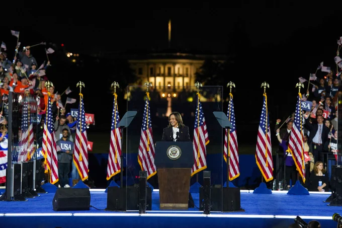 Photo of Vice President Kamala Harris delivering her closing remarks on a stage at the Ellipse in Washington, D.C. There is a crowd cheering on each side, with American flags behind her, and the White House dimly lit in the background. 