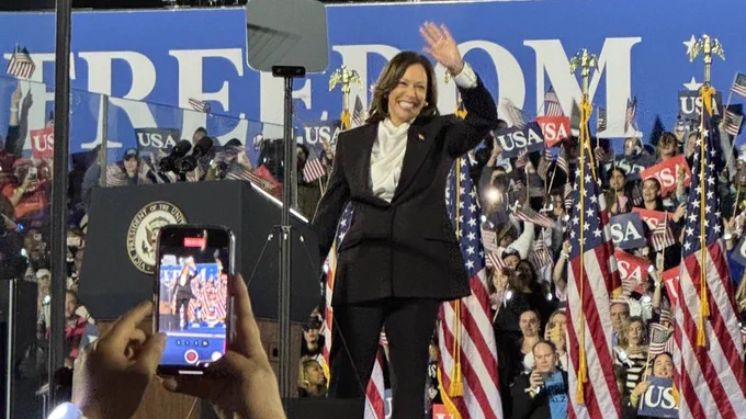 Photo of Vice President Kamala Harris waving on stage. American flags line the stage behind her. There’s a large banner that says “Freedom” hanging behind her.