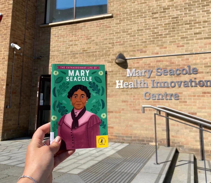 Image shows someone holding a book about Mary Seacole in front of the Mary Seacole Health Innovation Centre