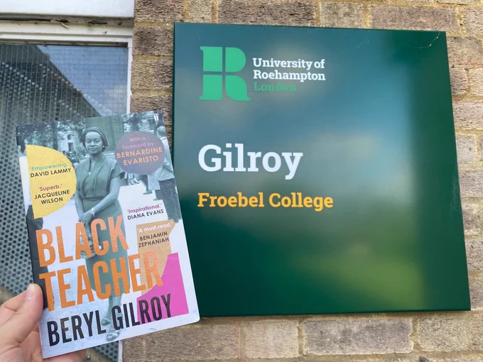 Image shows someone holding the book Black Teacher by Beryl Gilroy in front of a sign for the Gilroy building 