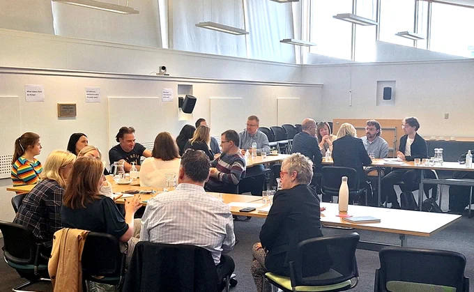 A photo taken at an in-person meeting of the CCPS Comms Member Advisory Group in October 2024, hosted at Quarriers Conference Centre in Glasgow. Around 20 people sit around tables in a large, brightly lit conference room, having discussions in groups of 4-5.