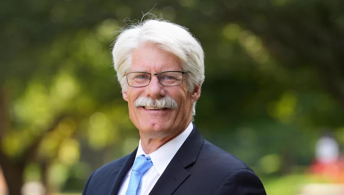 White man with white hair and mustache wears glasses and dark suit outdoors.