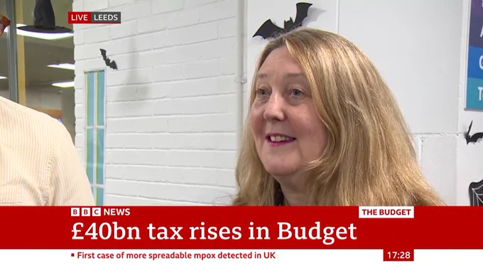 Good Things CEO, Helen Milner smiles during an interview with BBC News. There is a banner displaying headlines about £40bn tax rises in the budget and a case of spreadable mpox in the UK. The setting is indoors in a gym in Pudsey, Leeds.
