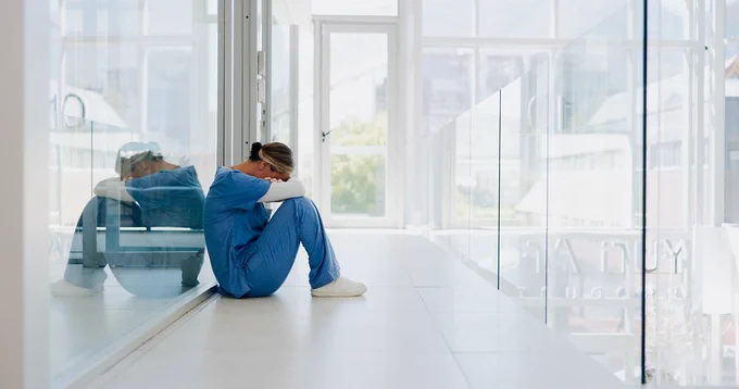 A distressed female healthcare worker sits on the floor of a hospital corridor with her head in her hands.