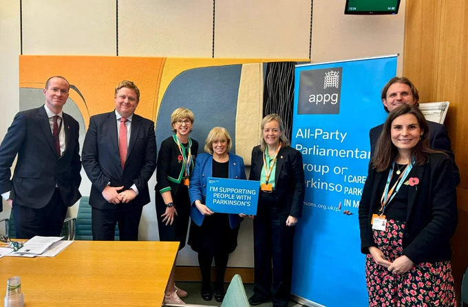 a group photo including members of Parliament and Parkinson's UK. There is a woman in the middle holding a blue placard which reads 'I'm supporting people with Parkinson's' and there is a bright blue banner which reads 'All-part Parliamentary group on Parkinson's.'