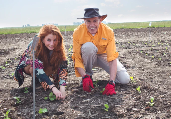 A red headed woman on the left and a man in a hat on the right work in a field with young green plants and brown dirt.