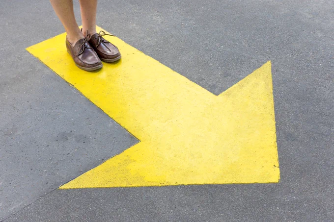 Brown shoes standing on a large yellow arrow painted on gray asphalt, pointing to the right, symbolizing movement and a new direction.