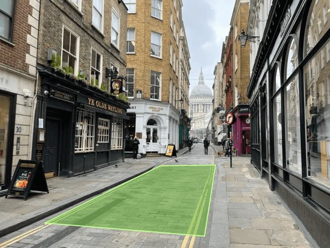 An image of a cobblestone street lined with businesses, including "Ye Olde Watling" pub, leading towards St. Paul's Cathedral in the distance. The street is mostly empty with a few pedestrians, and there is a distinctive green patch on the pavement.