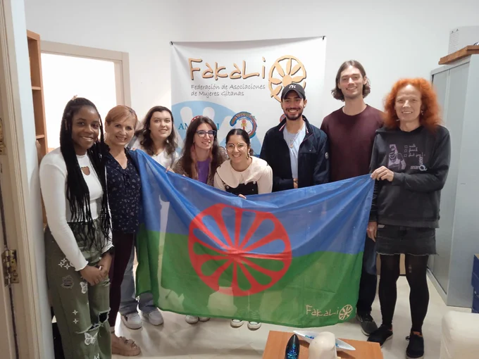Students taking a group picture with members of the Romani's Women's Association. Two individuals are holding the organization's flag.