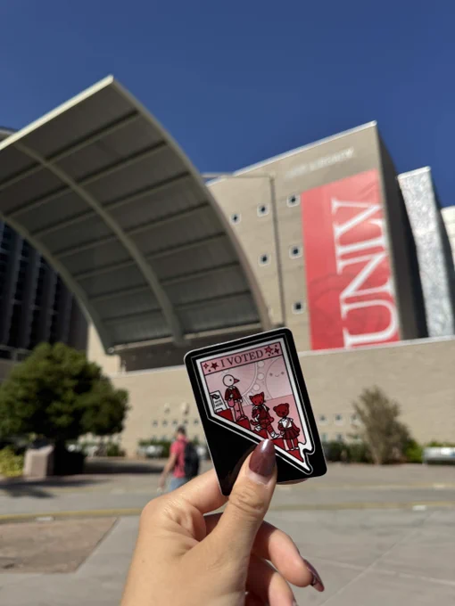 An I Voted sticker held up in front of a tall library building with a red UNLV banner