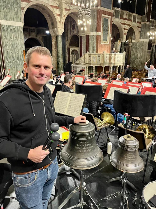 Olly Yates posing with 2 Forever Bells during rehearsal in Westminster Cathedral. David Hill conducts in the background