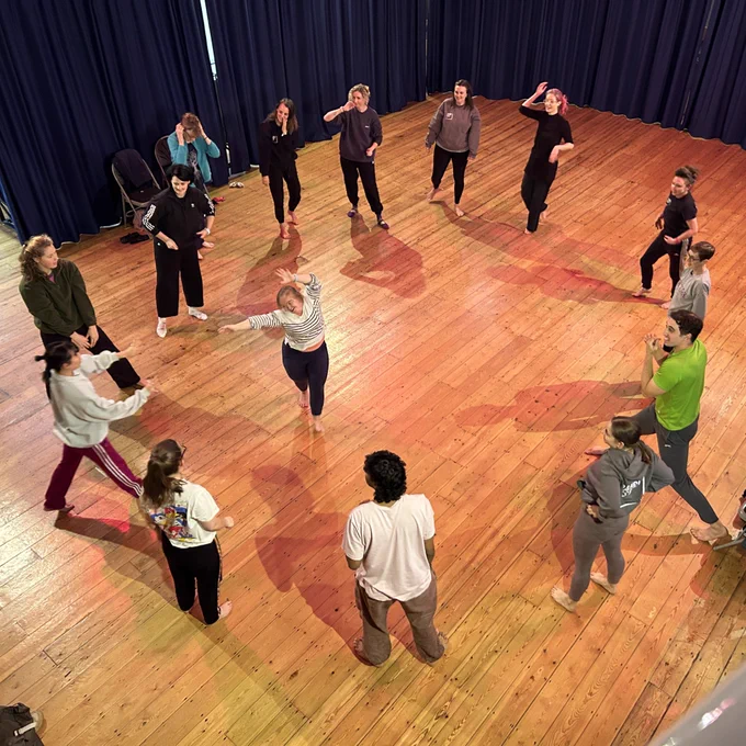 A dance workshop photo - taken from above - a group of adults all in a circle, with one dance in the middle reaching out.