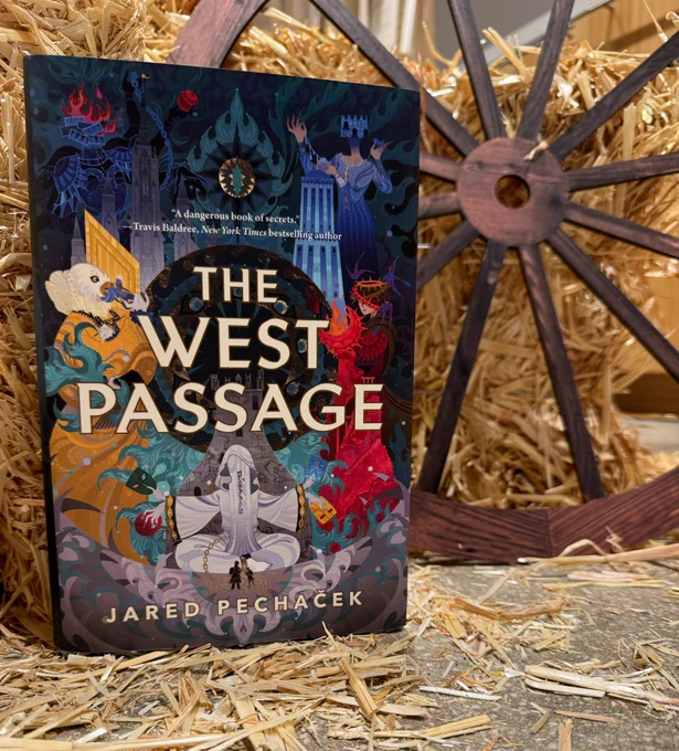 A copy of The West Passage by Jared Pechaček standing in front of a bunch of straw and a wooden wheel.