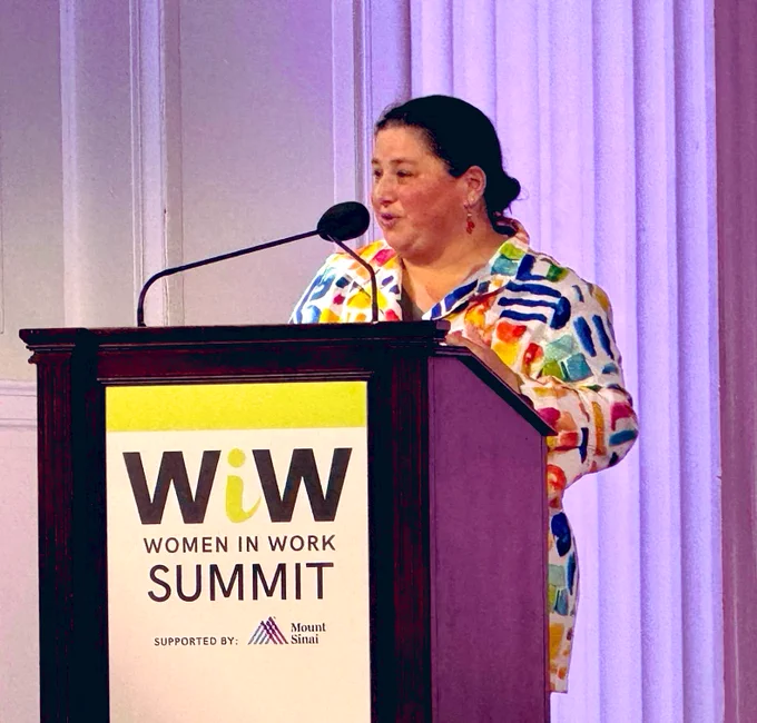 White woman with black hair wearing multi-colored jacket standing at a podium in front of lit wall.  Sign on podum says WiW women in work summit.
