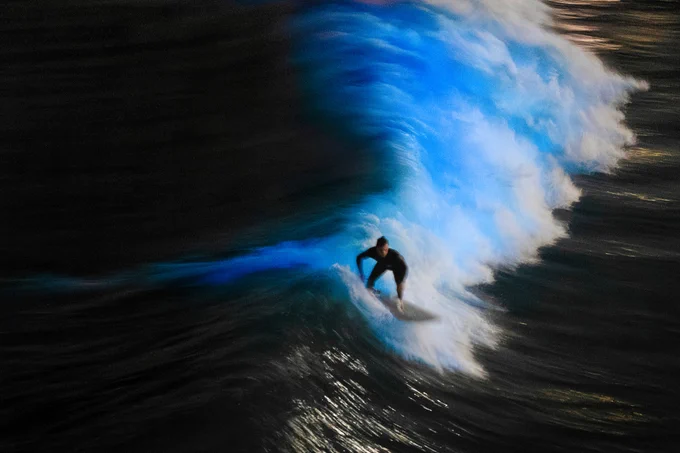 A person surfing a large wave at night with bioluminescence, captured with a motion blur effect.