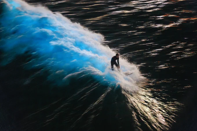 A person surfing a large wave at night, illuminated by bioluminescence on the water.
