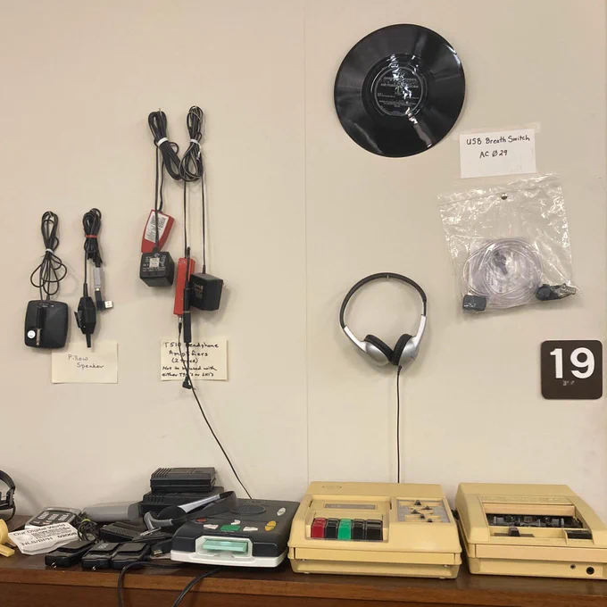 A beige wall with several assistive technology devices for listening to audiobooks, including headphones, cords, and cassette players.