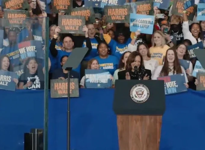 Kamala Harris speaking at a podium with people cheering and holding signs in the background, including Teamsters Against Trump supporters from Local 391 wearing Teamsters for Harris t-shirts.