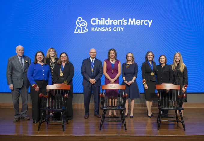 Group of eight individuals standing on a stage with chairs and floral arrangements in front of a backdrop displaying the Children's Mercy Kansas City logo.