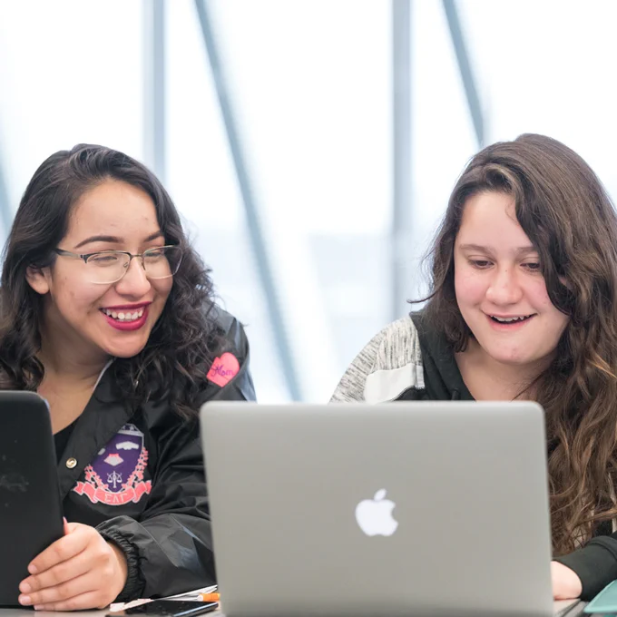 A photo of two people smiling, sitting at laptop computers.  