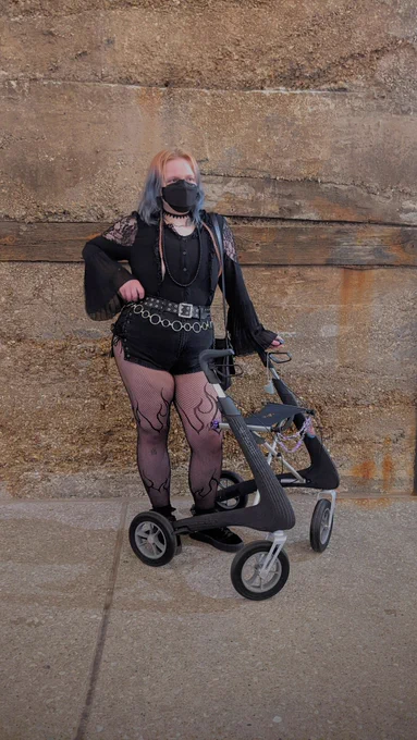 Jules, a white nonbinary person with medium length blue hair with two long pink braids, wearing a black mask with an all black outfit and standing with their dark grey rollator. They have extremely short shorts that tie up on the sides, a black button down with flowy sleeves and lacy shoulders, layered belts, and tights with flames on them.