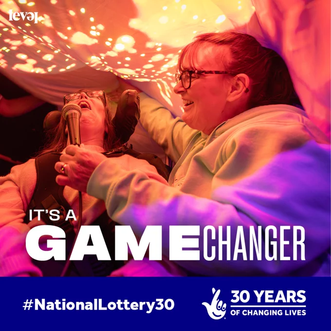 Two people sit under some fabric which is lit with sparkly colourful patterns in pink, orange and purple. One person holds a microphone for the other, who is a wheelchair user. They are both laughing and smiling and having fun. White text reads It's a gamechanger. The National Lottery logo sits below with white text that reads 30 years of changing lives.