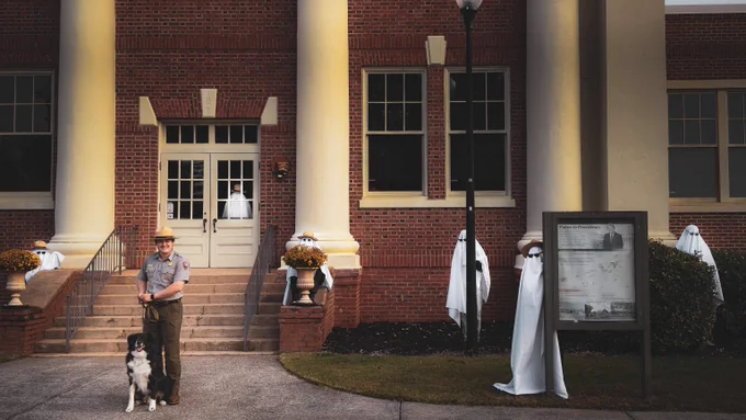 A National Park Ranger stands in front of the Plains High School front steps. Beside him is a fluffy, large, black and white dog. In the background, ghost (park rangers in white sheets) are hiding in bushes and behind objects. 