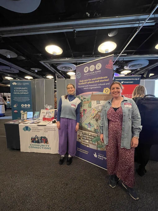 Rosie and Erin stand next to a promotional Make Your Mark banner that says "help care for and celebrate Scotland's places and stories!".