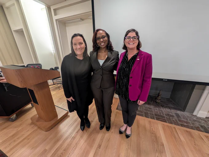 Emily, Dr. Warren and Leslie smile at the camera with their arms around each other next to a lectern in Marie Mount Hall